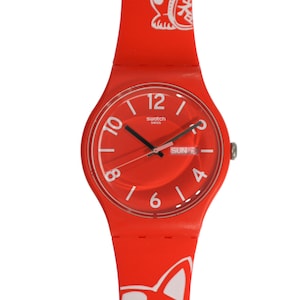 Könnte beinhalten: Eine rote Swatch-Uhr mit einem weißen Katzenmotiv auf dem Armband. Das Zifferblatt der Uhr ist rot mit weißen Zahlen und Zeigern. Die Uhr hat ein Datumsfenster, das "SUN 7" anzeigt.
