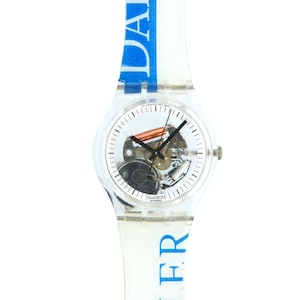 Può includere: Un orologio Swatch in plastica trasparente con quadrante bianco e cinturino a fantasia blu e bianca. L'orologio ha un movimento scheletrato, il che significa che il meccanismo interno dell'orologio è visibile. Il cinturino ha le parole "Daily" e "Sister" stampate su di esso.