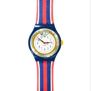 Könnte beinhalten: Eine blaue und goldene Swatch-Uhr mit weißem Zifferblatt und rot-weiß-blau gestreiftem Armband.