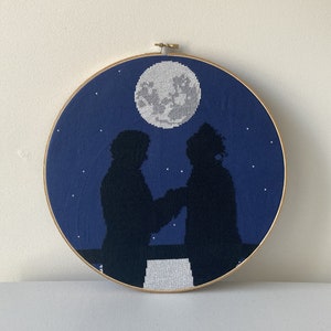 以下が含まれることがあります： 満月と星空の背景に、2人のシルエットが立っている刺繍のフープアートです。