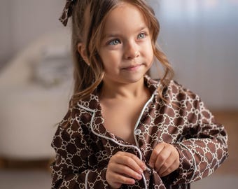 May include: A young girl wearing brown pajamas with a white geometric pattern and white trim. She has a matching bow in her hair. The pajamas are button-down and appear to be made of a soft material.