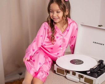 May include: A young person wearing pink pajamas with heart patterns and a matching bow. The pajamas are paired with a white record player with the brand name "CROSLEY" visible on the front. The record player is on a neutral-toned surface.
