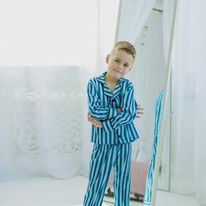 Op de afbeelding: Een kind draagt een blauw en teal gestreepte pyjamaset. De pyjamatop heeft een knoopsluiting en lange mouwen. De broek is lang en heeft een bijpassend streepjespatroon. Het kind staat voor een spiegel.