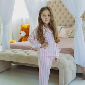 Könnte beinhalten: Ein junger Mensch trägt einen rosa-weiß gestreiften Pyjama mit Federbesatz. Der Pyjama besteht aus einem Hemd mit Knöpfen und einer Hose. Die Person steht vor einem Bett mit einem Teddybär und dekorativen Vorhängen.