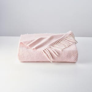 Puede incluir: Una manta de lana rosa suave con flecos blancos. La manta está doblada por la mitad y tiene un patrón de espiga.