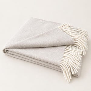 May include: A folded light beige throw blanket with a subtle woven texture. The blanket features long, cream-colored fringe detailing along the edges, providing a soft and tactile finish to the overall design of the textile piece.
