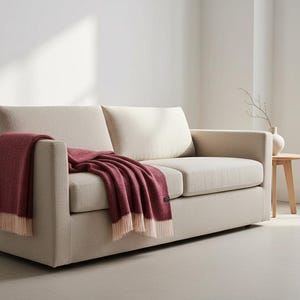 A dark red wine colour blanket made of wool draped from a light beige sofa in a light Scandinavian style room