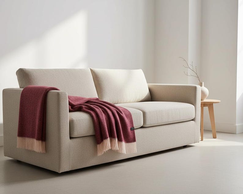 A dark red wine colour blanket made of wool draped from a light beige sofa in a light Scandinavian style room