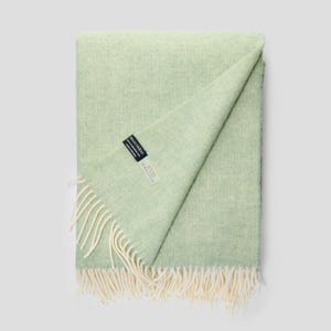 Peut inclure: Une couverture vert clair avec un motif de tissage en chevrons. La couverture a une frange crème sur un bord et une étiquette "moo ridine". Elle est pliée, avec un coin rabattu, révélant le dessous.