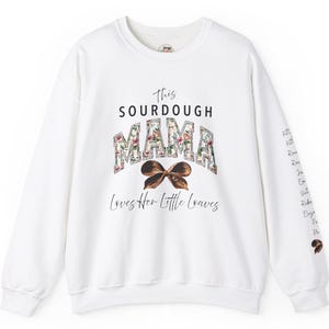 Può includere: Felpa bianca con un design floreale che dice "This Sourdough Mama Loves Her Little Loaves". Il design floreale è sulla parola "Mama" e sotto c'è un fiocco marrone. La manica ha un elenco di nomi scritti verticalmente.