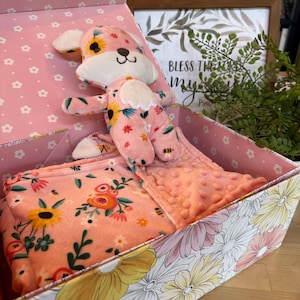 May include: A pink floral-patterned gift box contains a plush dog toy and a matching blanket. The dog toy has sunflower accents, and the blanket has a soft, textured surface. The box is decorated with daisy patterns.