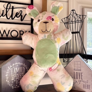 May include: A plush bunny-bear hybrid toy with a floral pattern in pink, green, and yellow. The toy has a green belly patch and a smiling face. Decorative signs with text are in the background.