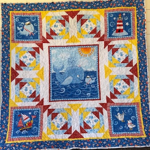 May include: A blue and yellow patchwork quilt with a nautical theme. The center panel features a whale and a smaller whale with a sailor hat. The surrounding panels feature lighthouses, sailboats, and seagulls.