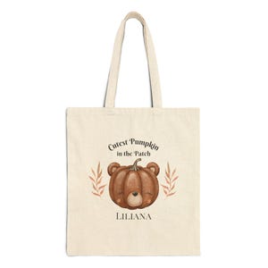 Könnte beinhalten: Eine beige Tragetasche mit einem braunen Kürbis mit einem Bärenkopf. Der Text auf der Tasche lautet "Cutest Pumpkin in the Patch" und "Liliana".