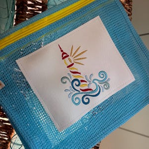 May include: A light blue mesh zippered pouch with a white fabric patch featuring an embroidered lighthouse with yellow, red, and blue details. The lighthouse is surrounded by blue and white waves.