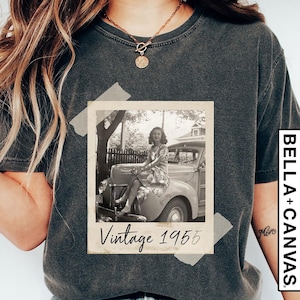 May include: Dark gray t-shirt featuring a vintage photograph of a woman sitting on a classic car. The photo is labeled "Vintage 1955" and is taped to the shirt. The shirt has a Bella+Canvas label.