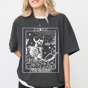 May include: A black t-shirt with a white graphic of a tarot card depicting a skeleton reading a book. The card is titled "The Reader".