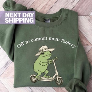 May include: Military green sweatshirt with the phrase "Off to commit mom foolery" in white. The graphic features a green frog wearing a hat and riding a scooter. The sweatshirt is a solid color.