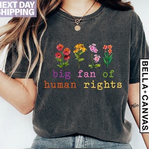 May include: Dark grey heather t-shirt with the text "big fan of human rights" in purple and orange. The shirt features embroidered flowers in red, yellow, pink, and orange. The shirt is a Bella+Canvas brand.