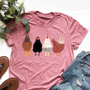 Peut inclure: Un t-shirt rose avec un motif de quatre poulets en rang. Les poulets sont bruns, noirs, blancs et brun et blanc.