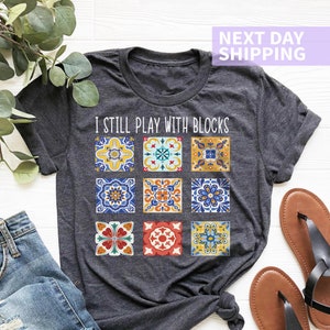 May include: A dark gray t-shirt with the text "I still play with blocks" printed on the front. The text is surrounded by a colorful pattern of nine square tiles with intricate designs.