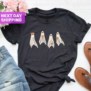 以下が含まれることがあります： 4体の幽霊が花を持っているグラフィックがプリントされた黒いTシャツ。幽霊は白で、目は黒。花はオレンジと黄色です。