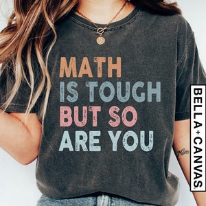 Op de afbeelding: Een donkergrijs T-shirt met de tekst "MATH IS TOUGH BUT SO ARE YOU" in kleurrijke, verweerde letters.