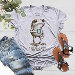 May include: Athletic heather gray t-shirt featuring a detailed illustration of a frog holding a mug. The text "I HAVE TO SAY WEIRD STUFF OR I'LL DIE" is printed below the frog. The shirt is paired with blue jeans and brown sandals.