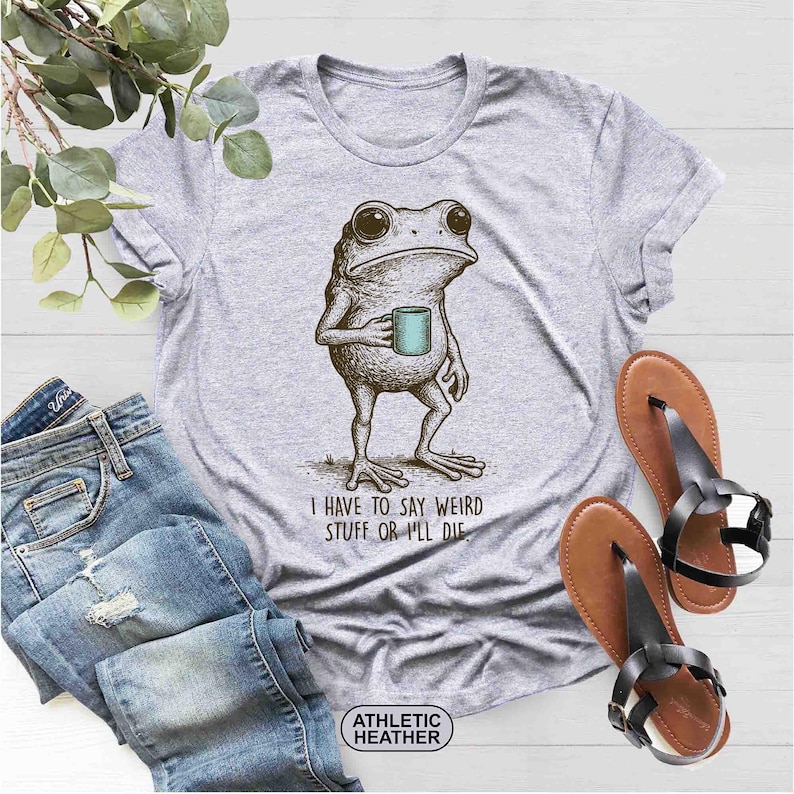 May include: Athletic heather gray t-shirt featuring a detailed illustration of a frog holding a mug. The text "I HAVE TO SAY WEIRD STUFF OR I'LL DIE" is printed below the frog. The shirt is paired with blue jeans and brown sandals.
