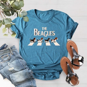K&ouml;nnte beinhalten: Ein t&uuml;rkisfarbenes T-Shirt mit einem Aufdruck von vier Beagles, die &uuml;ber einen Zebrastreifen laufen. Der Text "THE BEAGLES" befindet sich &uuml;ber den Beagles.