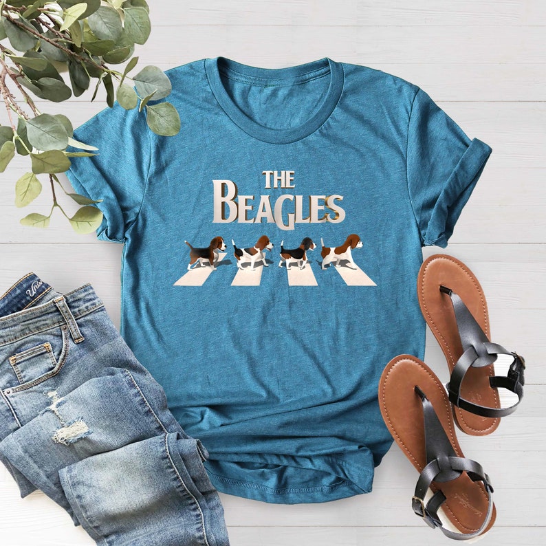 K&ouml;nnte beinhalten: Ein t&uuml;rkisfarbenes T-Shirt mit einem Aufdruck von vier Beagles, die &uuml;ber einen Zebrastreifen laufen. Der Text "THE BEAGLES" befindet sich &uuml;ber den Beagles.