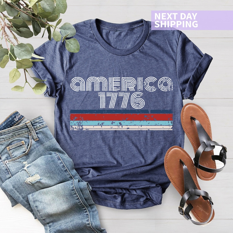 May include: A navy blue t-shirt with a white graphic that says "America 1776" in a retro font. The graphic is layered with red, blue, and white stripes.