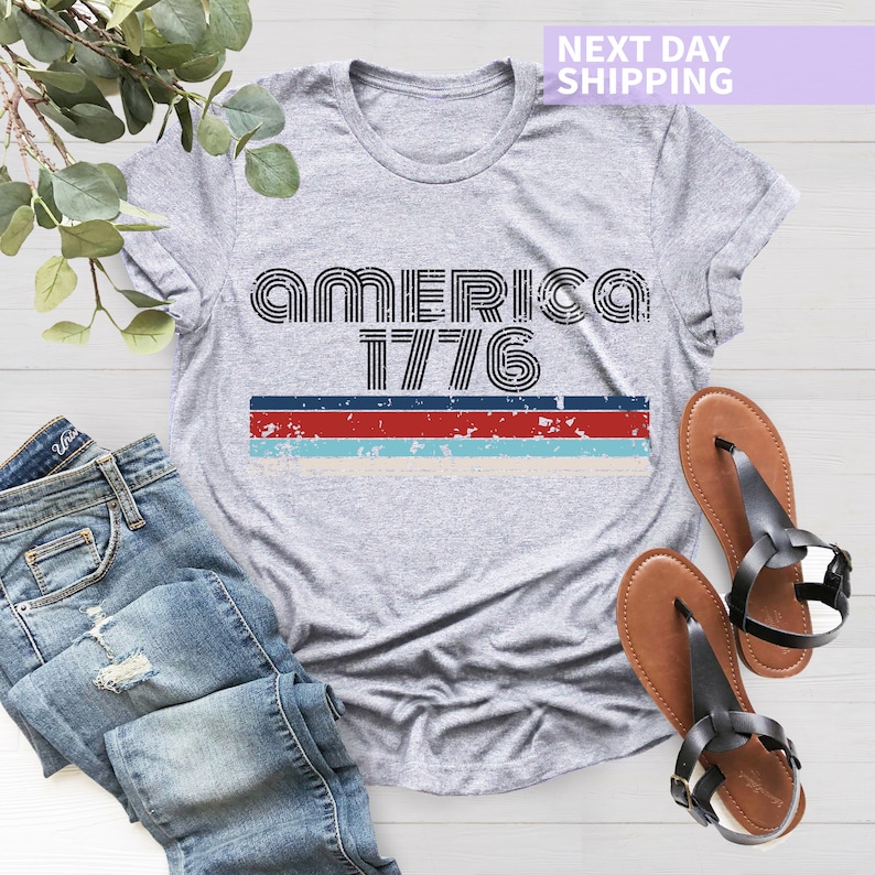 May include: A gray t-shirt with a distressed, vintage-style graphic that reads "America 1776". The graphic features red, white, and blue stripes.