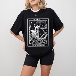 May include: Black t-shirt with a white graphic of a skeleton reading a book. The graphic is surrounded by flowers and stars. The text "THE READER" is below the graphic.