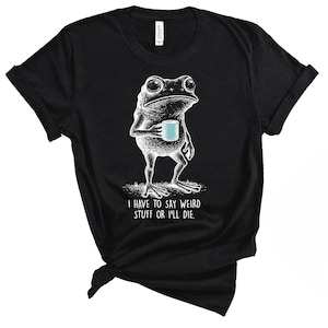 May include: Black t-shirt featuring a white illustration of a frog holding a blue mug. The frog has a contemplative expression. The text "I HAVE TO SAY WEIRD STUFF OR I'LL DIE" is below the frog. The shirt is made of a soft material.