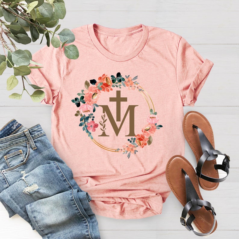 May include: A light pink t-shirt featuring a floral wreath design with a cross and the letters "M" and "T". The wreath is filled with pink, peach, and green flowers. A pair of brown sandals with black straps and blue jeans are also in the image.