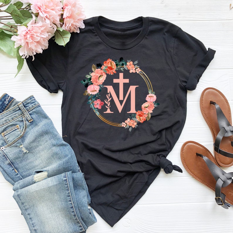 May include: Black t-shirt featuring a floral wreath design with a cross and the letter "M" in a light pink color. The wreath includes pink and orange flowers and green leaves. The shirt is paired with blue jeans and brown sandals.