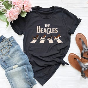 K&ouml;nnte beinhalten: Schwarzes T-Shirt mit einem Grafikdruck von vier Beagles, die &uuml;ber einen Zebrastreifen laufen. Der Text "THE BEAGLES" befindet sich &uuml;ber den Beagles.
