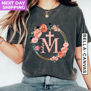 May include: Dark gray t-shirt featuring a floral wreath design with pink flowers and the letters "M" and a cross. The design is set within a gold-colored circular frame. The shirt is from Bella+Canvas.