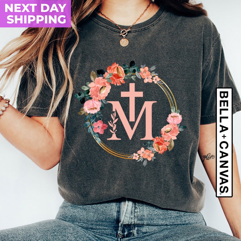 May include: Dark gray t-shirt featuring a floral wreath design with pink flowers and the letters "M" and a cross. The design is set within a gold-colored circular frame. The shirt is from Bella+Canvas.