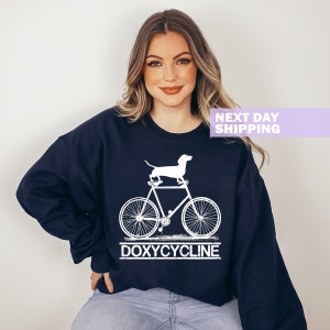 May include: A navy blue sweatshirt with a white graphic of a dachshund riding a bicycle. The text "DOXYCYCLINE" is printed below the bicycle.