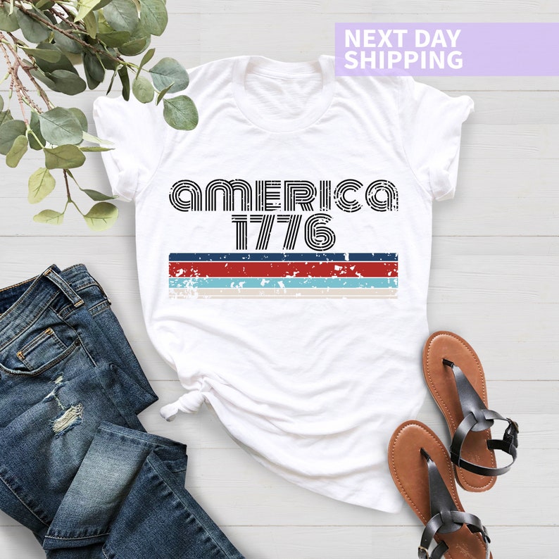 May include: White t-shirt with a black graphic that says "AMERICA 1776" with red, white, and blue stripes underneath.