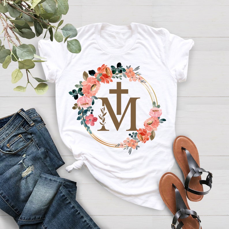 May include: White t-shirt featuring a floral wreath design with a cross and the letters "M". The wreath includes pink and orange flowers and green leaves. The shirt is paired with blue jeans and brown sandals.