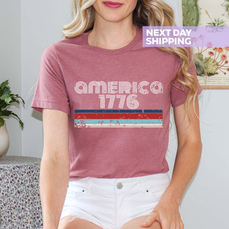 May include: A maroon t-shirt with a white graphic that says "AMERICA 1776" in a retro font. The graphic is layered with red, white, and blue stripes.