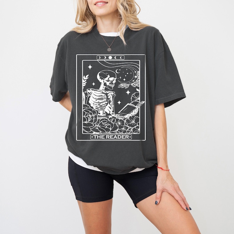 May include: A black t-shirt with a white graphic of a tarot card depicting a skeleton reading a book. The card is titled "The Reader".