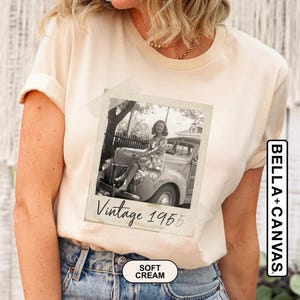 May include: Soft cream-colored t-shirt featuring a vintage black and white photo of a woman sitting on a classic car. The photo is labeled "Vintage 1955." The shirt is made of soft material and has the "BELLA+CANVAS" logo.