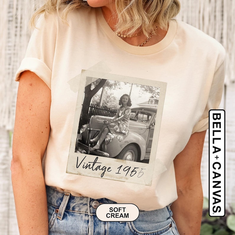 May include: Soft cream-colored t-shirt featuring a vintage black and white photo of a woman sitting on a classic car. The photo is labeled "Vintage 1955." The shirt is made of soft material and has the "BELLA+CANVAS" logo.
