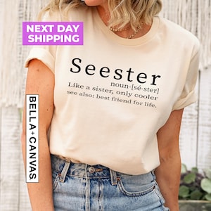 Pu&ograve; includere: Una maglietta beige con testo nero che recita "Seester noun-[s&eacute;-ster] Like a sister, only cooler see also: best friend for life." La maglietta &egrave; indossata da una persona con jeans blu.