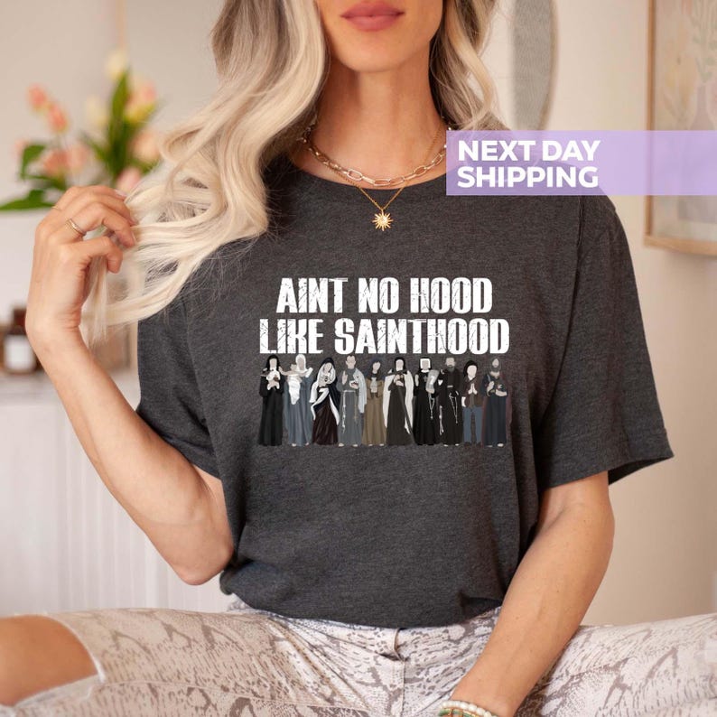 K&ouml;nnte beinhalten: Ein dunkelgraues T-Shirt mit dem Text "Ain't no hood like Sainthood" in wei&szlig; gedruckt. Das Shirt zeigt eine Grafik einer Gruppe von Menschen in wei&szlig;en Roben.