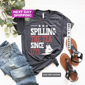 Pu&ograve; includere: Maglietta color grigio scuro melange con il testo "Spilling the Tea Since 1773" in rosso e bianco. Una silhouette bianca di una nave si trova sotto il testo. La maglietta &egrave; stesa su una superficie bianca.